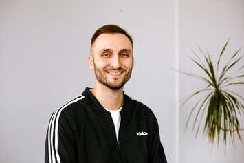 Ein junger Mann mit Dreitagebart und kurzen schwarzen Haaren lächelt in die Kamera. Er trägt eine schwarze Adidas-Jacke mit weißen Streifen an den Armen.