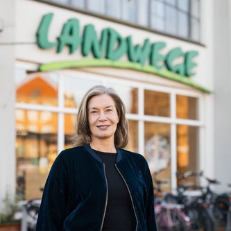 Eine Frau mit grau-blonden, halblangen Haaren steht lächelnd vor einem Gebäude mit der Aufschrift „LANDWEGE“. Im Hintergrund sind Fahrräder und große Fenster zu sehen. Die Umgebung wirkt städtisch und belebt.