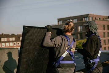 Zwei Personen stehen mit dem Rücken zur Kamera auf einem Dach und halten ein PV-Paneel in der Hand. Sie tragen lilafarbenes Sicherheitsgeschirr. Die Sonne scheint.