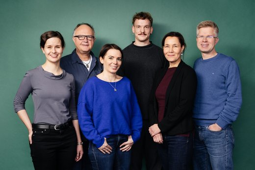 Eine Gruppe von sechs Personen posiert lächelnd vor einem grünen Hintergrund. Sie stehen eng beieinander und tragen legere Kleidung. Die Atmosphäre wirkt freundlich und entspannt.