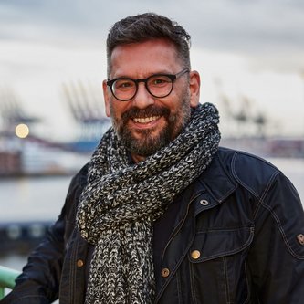Ein Mann mit Bart und Brille lächelt in die Kamera. Er trägt einen dunkelgrauen Schal und eine schwarze Jacke. Im Hintergrund ist ein Hafen mit Kränen zu sehen.