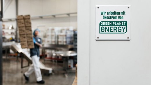 Eine Person trägt Kartons in einer Fabrikumgebung. An der Wand hängt ein Schild mit der Aufschrift: "Wir arbeiten mit Ökostrom von Green Planet Energy." Industrie, Nachhaltigkeit, erneuerbare Energie.