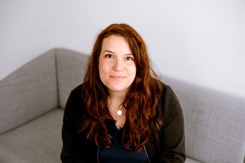 Eine Frau mit roten langen und gewellten Haaren schaut nach oben in die Kamera. Sie trägt dunkle Klamotten und eine dezente Silberkette.