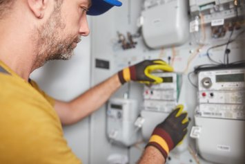 Ein Mann mit Bart in gelben T-Shirt und mit Cappi arbeitet an einem Zählerkasten. Er trägt Arbeitshandschuhe und schaut konzentriert auf die Geräte an der Wand.