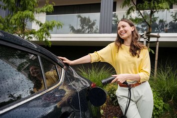Eine Frau mit braunen langen Haaren und gelben Shirt steht vor einem begrünten Haus an einem schwarzen Auto und hält einen Ladestecker in der Hand.