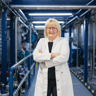 Eine lächelnde Frau in einem Laborkittel steht mit verschränkten Armen in einer industriellen Anlage mit Rohren und Maschinen. Die Umgebung ist hell beleuchtet.