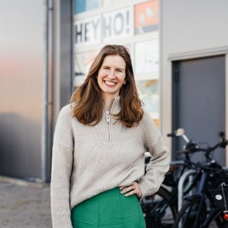 Eine Frau steht lächelnd vor einem Gebäude mit Fahrrädern im Hintergrund. An der Wand ist Schriftzug "HEY HO!". Moderne, urbane Umgebung sorgt für ein freundliches, einladendes Ambiente.