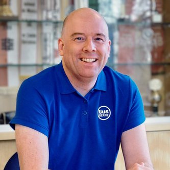 Eine lächelnde Person in einem blauen "tus BERNE"-Polo sitzt an einem Tisch. Im Hintergrund sind Regale mit Pokalen und Urkunden sichtbar.