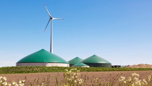 Eine Biogasanlage hinter einem Ackerfeld. Dahinter steht eine Windkraftanlage.