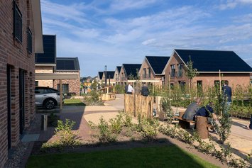 Moderne Reihenhäuser mit Solardächern säumen eine gepflegte Wohnanlage. Mehrere Personen unterhalten sich entspannt auf einer Bank. Der sonnige Tag unterstreicht die umweltfreundliche Architektur und das grüne Design.