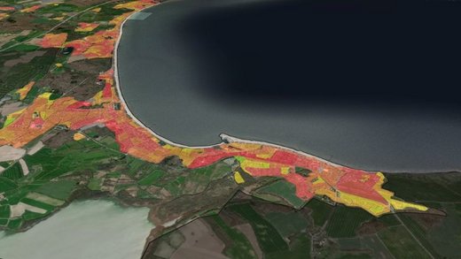 Luftaufnahme zeigt farbcodierte Landflächen nahe einer Küstenlinie. Die Flächen sind in Rot, Gelb und Grün markiert. Im Hintergrund ist ein großes Gewässer sichtbar. Text: "powered by greenventory."