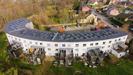 Geschwungenes Wohngebäude mit zahlreichen Photovoltaikmodulen auf dem Flachdach. Vor den Wohnungen befinden sich Balkone und Gärten, dahinter liegt eine Wohngegend mit weiteren Häusern und Bäumen.