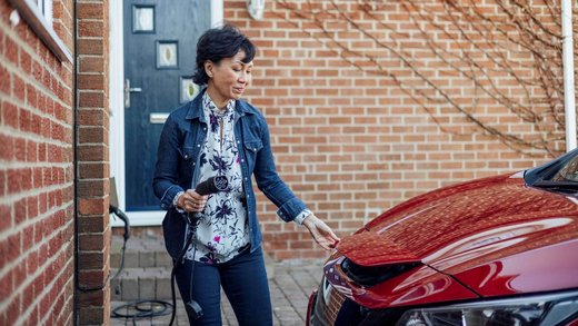 Eine Frau mit kurzen schwarzen Haaren und geblümter Bluse steht vor einem Backsteinhaus. Sie hält ein Ladekabel in der Hand, das an einer Wallbox am Haus befestigt ist und ist dabei, dieses in ein rotes Auto zu stecken.