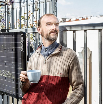 Ein Mann steht vor einem Balkonkraftwerk in der Sonne und hält eine weiße Tasse in der Hand. Er blickt an der Kamera vorbei und hat eine entspannte Körperhaltung.