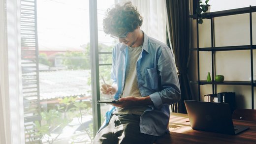 Junger Mann in grauem T-Shirt und offenem blauen Hemd lehnt an einem Schreibtisch und hält ein Tablet und einen Stift.