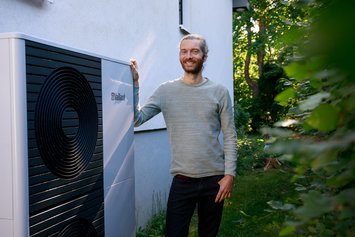 Ein Mann mit grauen Haaren und Zopf steht neben einem großen Kasten (Wärmepumpe). Im Hintergrund ist ein Haus und Garten zu sehen.