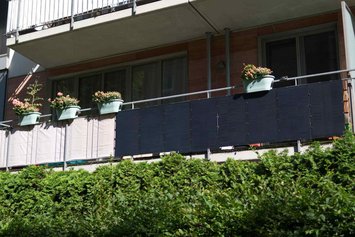 Ein Balkon mit blumig bepflanzten Pflanzkübeln und Balkonkraftwerk am Geländer. Die Sonne scheint.