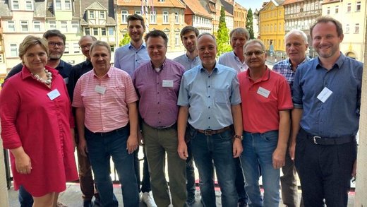 Eine Gruppe von zwölf Personen steht dicht zusammen auf einem Balkon. Sie lächeln und tragen Namensschilder. Im Hintergrund sind historische Gebäude und ein klarer Himmel zu sehen.