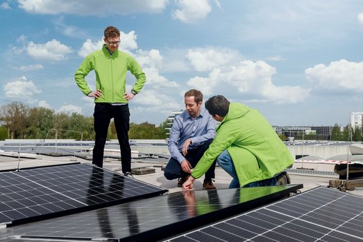 Drei Männer inspizieren Solarpaneele auf einem Flachdach bei klarem Himmel. Zwei tragen grüne Jacken, einer kniet und zeigt auf ein Paneel. Im Hintergrund sind Bäume und Gebäude sichtbar.