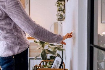 Eine Person in fliederfarbenem Pullover drückt auf einen Lichtschalter. Im Hintergrund sind Pflanzen und Regale zu sehen.