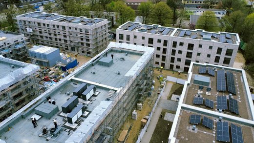 Mehrere Gebäude im Bau. Auf dem vorderen Flachdach stehen große Wärmepumpen und dort arbeiten mehrere Personen. Auf dem rechten und den dahinterliegenden Gebäuden sind Photovoltaikmodule in Reihen installiert.