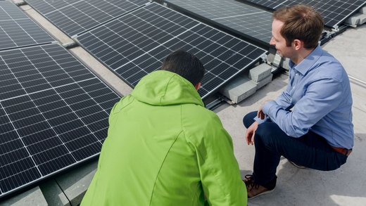 Ein Mann in grüner Jacke und ein Mann in blauem Hemd begutachten Solarpaneele auf einem Flachdach. Die Umgebung zeigt mehrere aufgereihte Solarmodule zur Energiegewinnung.