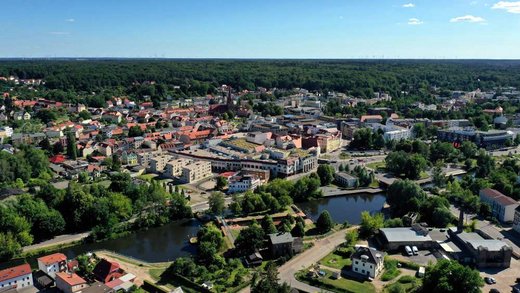 Luftaufnahme einer Stadt mit einer Vielzahl von Gebäuden und einem Fluss im Vordergrund. Die Umgebung ist von dichten Wäldern umgeben, die den Horizont begrenzen. Klarer Himmel.