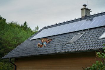 Lachende Familie, die aus dem Dachfenster in ihrem Haus mit PV-Module auf dem Dach schaut.