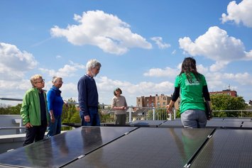 Eine Gruppe grauhaariger Menschen steht mit einer schwarzhaarigen Frau in grünem T-Shirt mit der Aufschrift "Green Planet Energy" auf einem Dach, das mit PV-Modulen bedeckt ist.