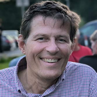 Ein Mann mit kariertem Hemd lächelt in die Kamera. Er sitzt im Freien, bei Abenddämmerung, mit unscharfen Personen und Autos im Hintergrund.