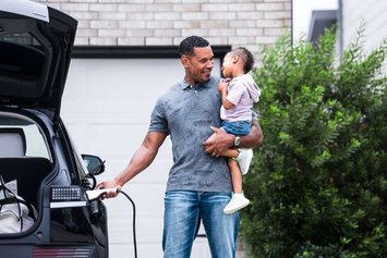 Ein Mann trägt ein kleines Kind auf dem Arm und lächelt es an, während er mit der anderen Hand ein E-Auto lädt.