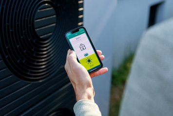 Kamerapersüektive über die Schulter einer Person in hellgrauem Pullover. Sie hält ein Smartphone in der Hand auf dem eine App eine Temperatur und Haus-Icon anzeigt. Im Hintergrund steht eine Wärmepumpe.