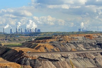 Braunkohletagebau im Vordergrund. Schwere Maschinen bewegen Erde. Im Hintergrund rauchende Industrieschornsteine vor bewaldetem Horizont. Wolken ziehen am Himmel entlang.