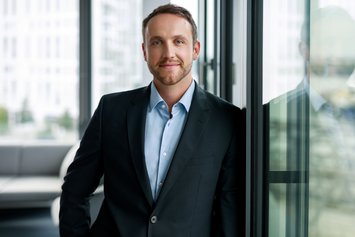 Ein Mann mit kurzen Haaren in hellblauem Hemd und dunklem Sakko lehnt in einem Büro an einer Fensterscheibe und lächelt leicht in die Kamera.