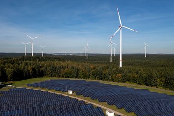 Windkraftanlagen und Solarpanels erzeugen erneuerbare Energie auf einer weitläufigen, bewaldeten Fläche unter klarem Himmel. Nachhaltigkeit und saubere Energie sind zentrale Themen in dieser Szene.
