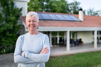 Eine Frau mit sehr kurzen weiß-grauen Haaren steht vor einem Haus mit PV-Anlage auf dem Dach. Sie trägt einen hellgrauen Pullover und lächelt mit verschränkten Armen in die Kamera.