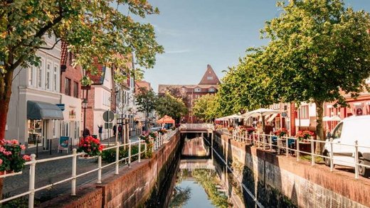 Straßenansicht: Ein ruhiger Kanal verläuft durch eine charmante historische Stadt mit Backsteinhäusern und Cafés, flankiert von Bäumen und Blumen. Der blaue Himmel spiegelt eine idyllische, friedliche Atmosphäre wider.