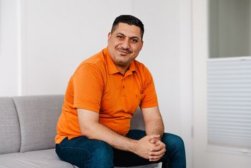 Ein Mann dunklen kurzen Haaren sitz auf einem hellgrauen Sofa und lächelt in die Kamera. Er hat seine Arme auf die Oberschenkel gestützt und die Hände zusammengelegt. Er trägt ein orangenes Polo-Shirt und eine dunkelblaue Jeans.