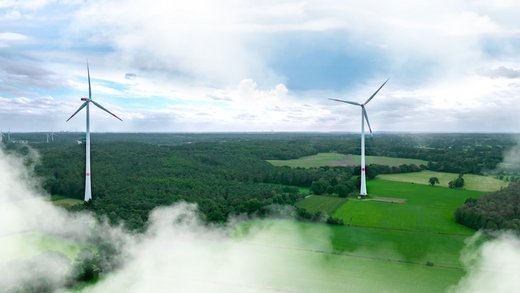 Zwei große Windräder drehen sich über grünen Feldern und Wäldern, umgeben von leichten Wolken, unter einem bewölkten Himmel. Erneuerbare Energie, Windkraft und Nachhaltigkeit stehen im Fokus.