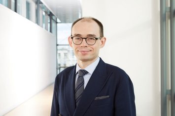 Ein Mann mit Brille trägt einen blauen Anzug mit Krawatte und steht in einem modernen, gut beleuchteten Innenflur mit Glasfenstern im Hintergrund.