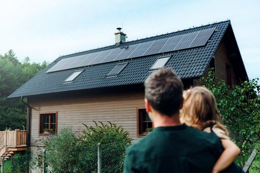 Ein Mann in dunkelgrünem Pullover trägt ein blondes Kind auf dem Arm und steht mit dem Rücken zur Kamera. Vor ihnen steht ein Holzhaus mit einer PV-Anlage auf dem Dach, das sie sich anschauen.