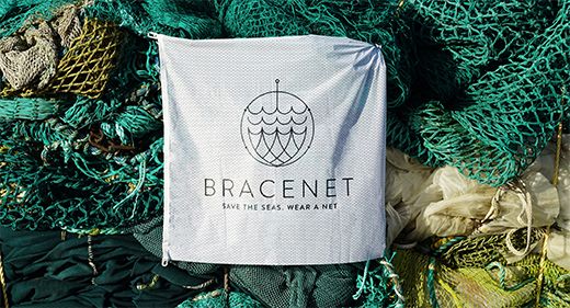 Ein weißes Banner mit dem Logo und Text "BRACENET - SAVE THE SEAS. WEAR A NET" hängt vor einem Stapel grüner Fischernetze im Freien. Thema: Umweltschutz, Recycling.