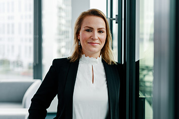 Eine blonde Frau in Geschäftskleidung steht in einem modernen Büro neben einer Glaswand. Im Hintergrund sind unscharf Büromöbel und Fenster zu erkennen. Die Frau lächelt leicht.